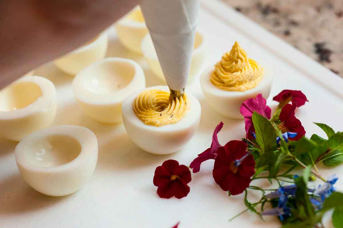 A pastry piping bag filling 2 deviled eggs on a cutting board next to garnish flowers.