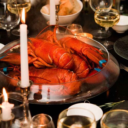 A table with a large bowl of cooked lobsters in the center, surrounded by glasses, candles and cutlery.