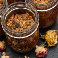 A jar of raspberry peach crisp on a table, sprinkled with a few small roses, beside 2 other jars.