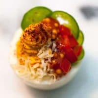 A deviled egg on a white plate garnished with jalapeño, cheese, peppers and spices.