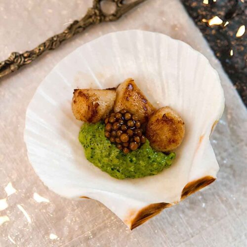 A small shell on a plate containing 3 small scallops, pesto cream and mustard caviar.