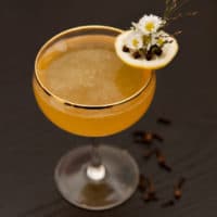 A cocktail on a table, garnished with a lemon slice, flowers and cloves, with a few cloves at the base.