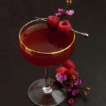 A cocktail on a table garnished with raspberries and small flowers, with small flowers and berries at its base.