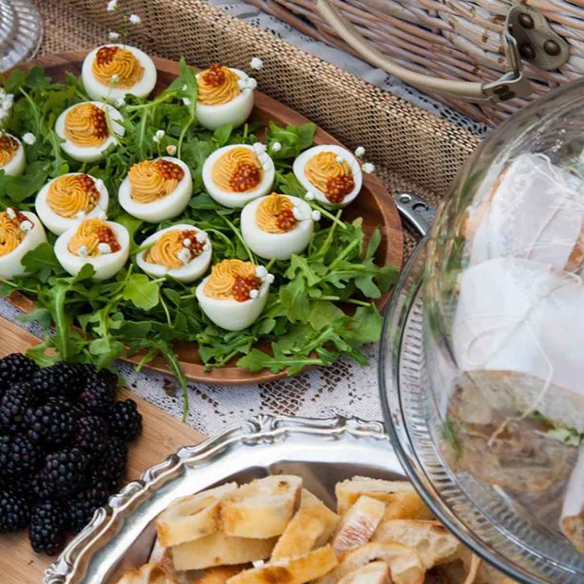 A picnic spread of fresh cut baguette, a plate of deviled eggs and sandwiches on a platter beside a picnic basket.