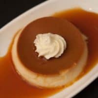 A flan on a plate, surrounded by caramel and topped with whipped cream.