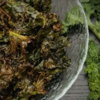 A glass bowl of kale chips on a table beside a few fresh leaves of kale.