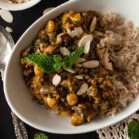 A bowl of curry, turkey butternut squash on a table with a lace table cloth, a bowl of shaved almonds and a few mint leaves.