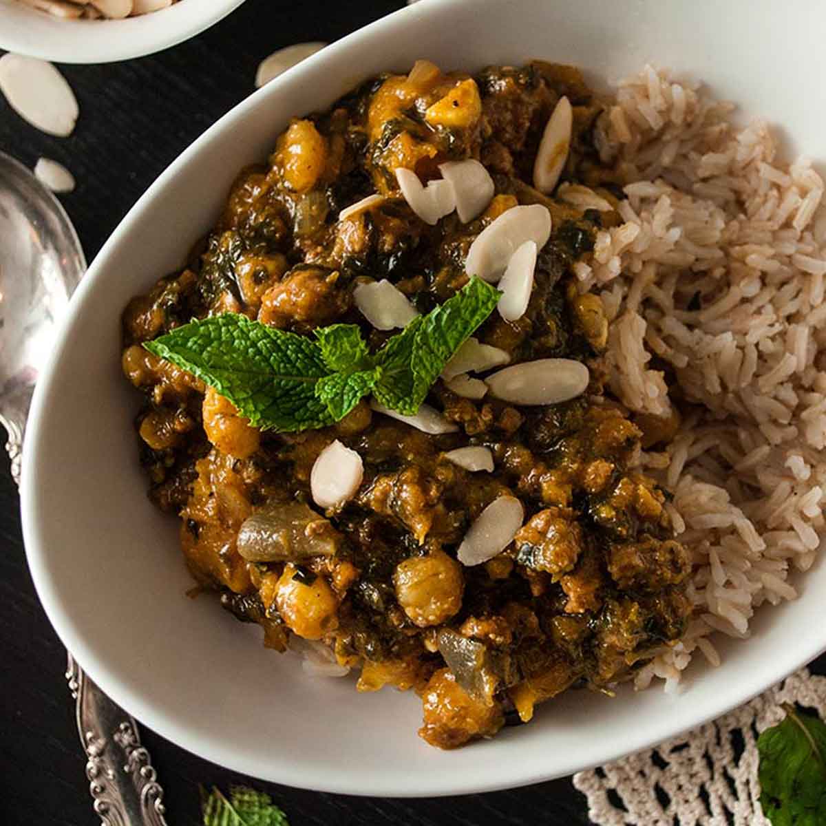 A bowl of curry, turkey butternut squash on a table with a lace table cloth, a bowl of shaved almonds and a few mint leaves.