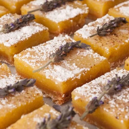 12 lavender lemon bars on a white marble table garnished with fresh lavender and powdered sugar.