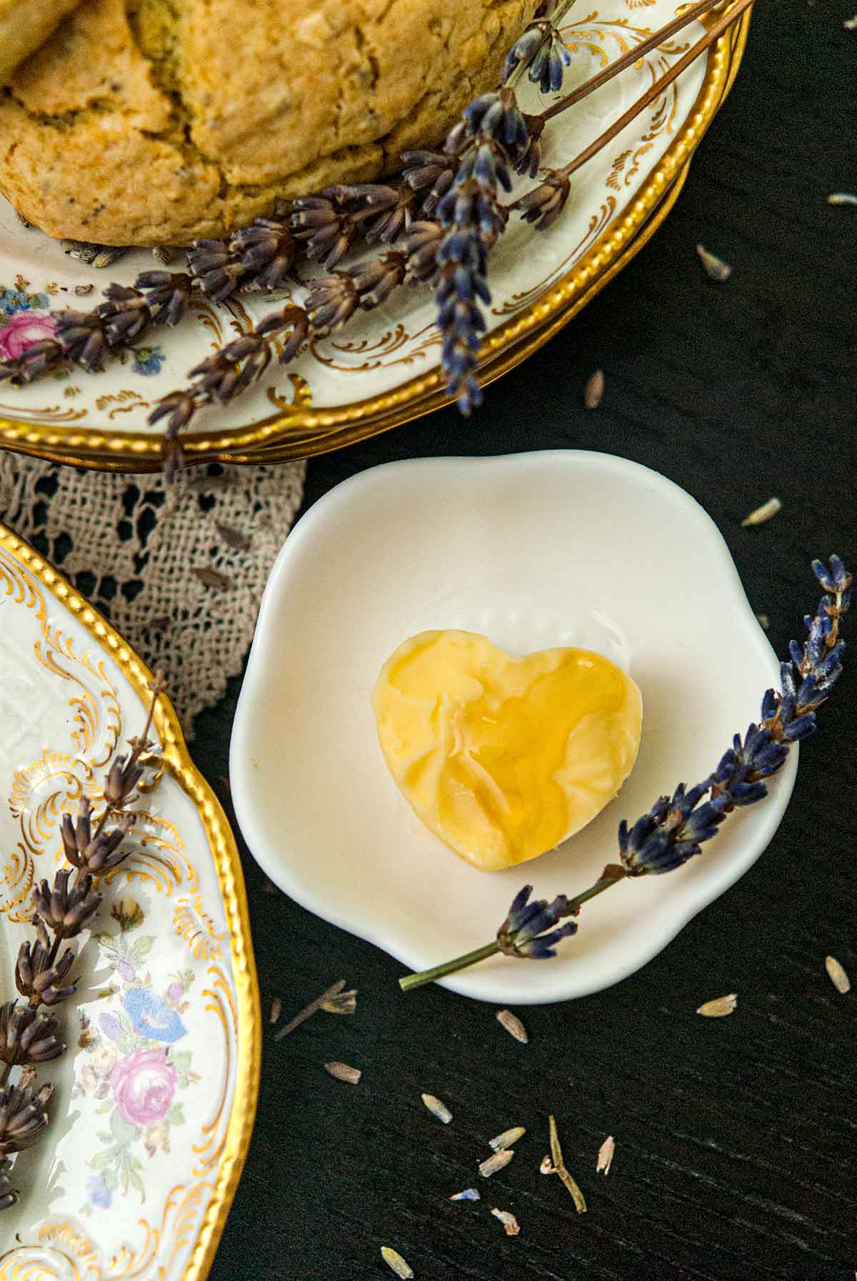 A small bowl with butter in the shape of a heart, garnished with honey and a sprig of lavender besides plates with lavender.