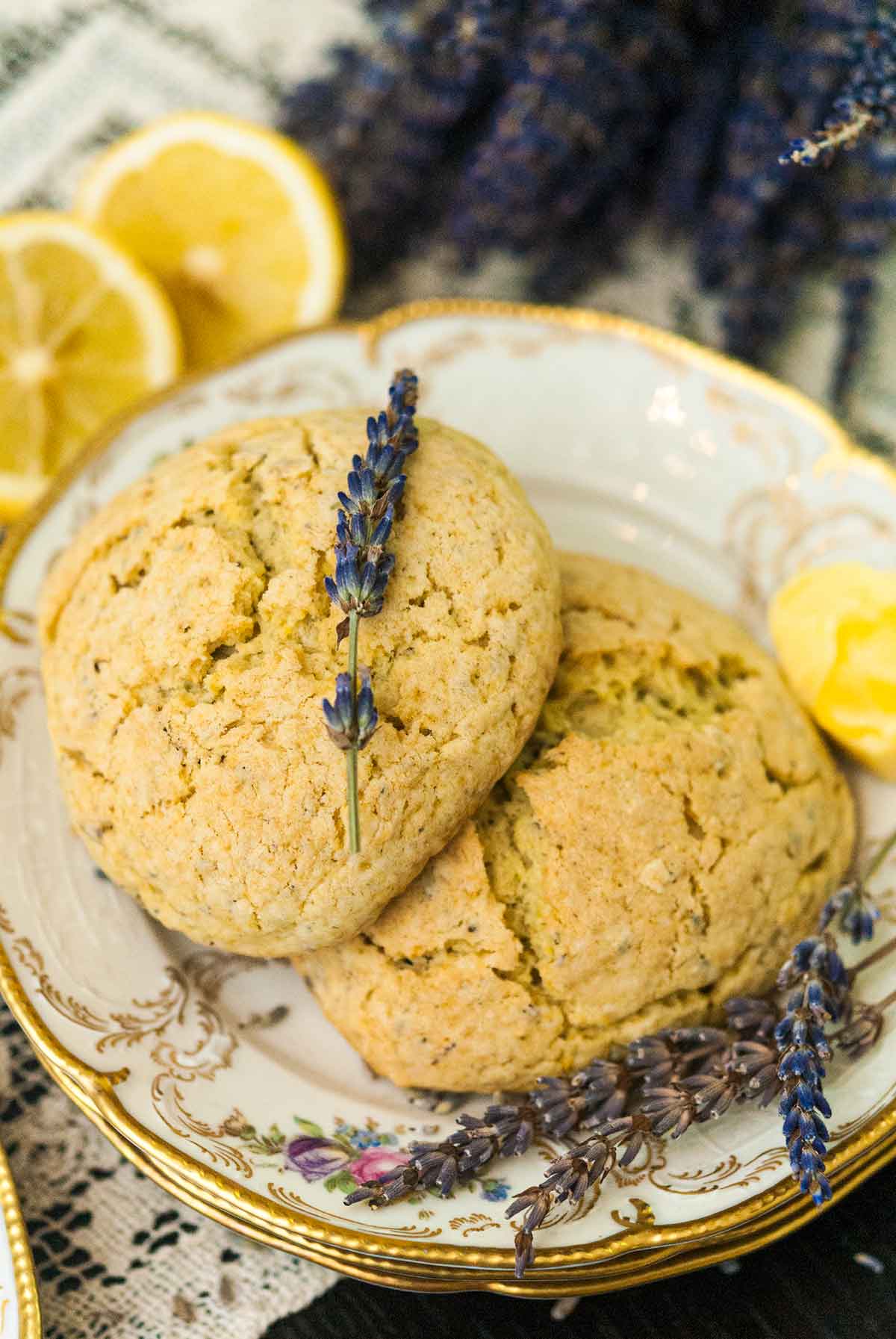 Two scones on plates with gold edges with fresh lavender placed on top, beside 2 cut lemons and a bunch of fresh lavender.