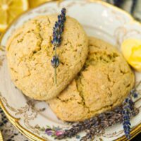 Two scones on plates with gold edges with fresh lavender placed on top, beside 2 cut lemons and a bunch of fresh lavender.