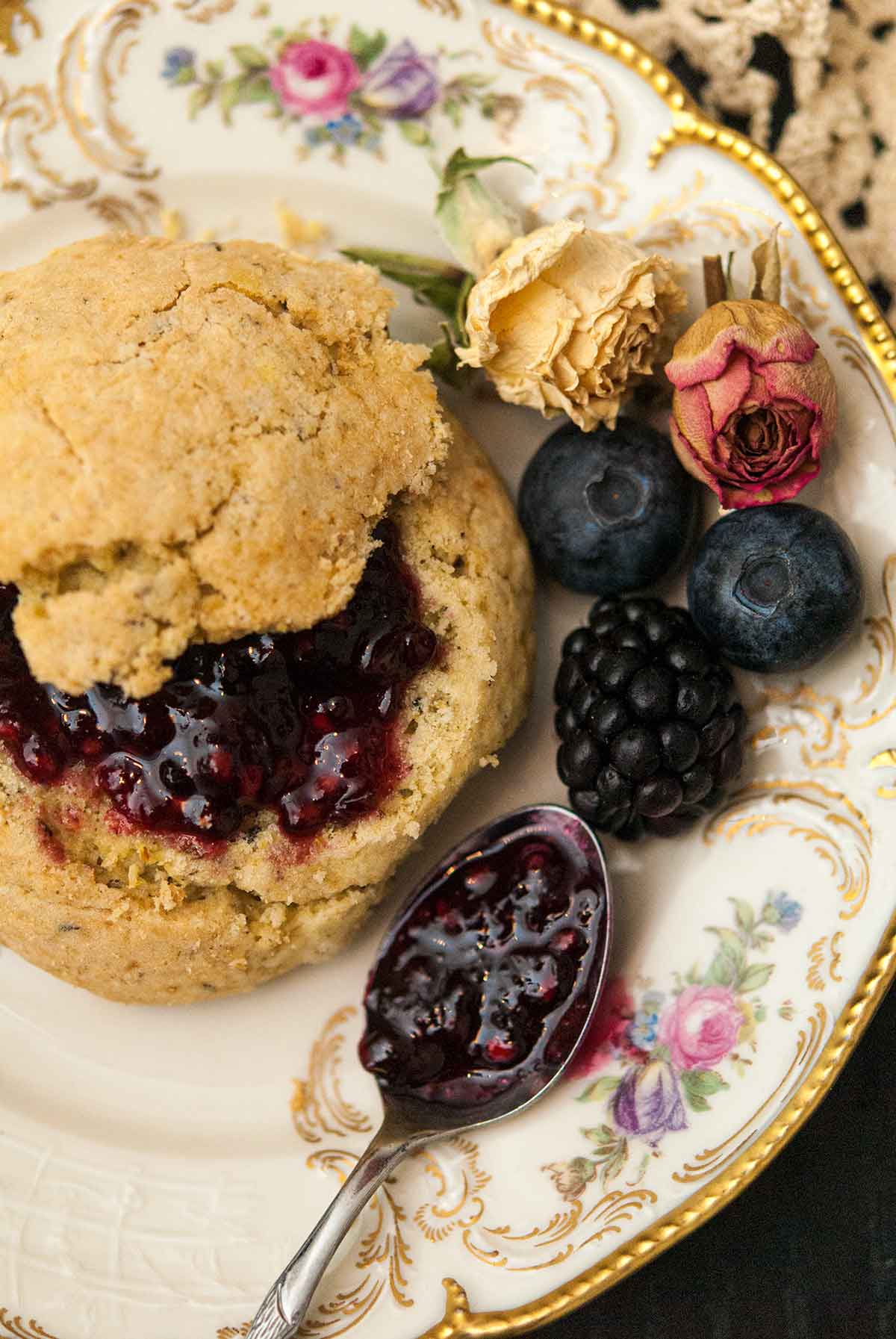 A scone with jam on a decorative plate next to a spoon with jam, a few berries and a few dry roses.