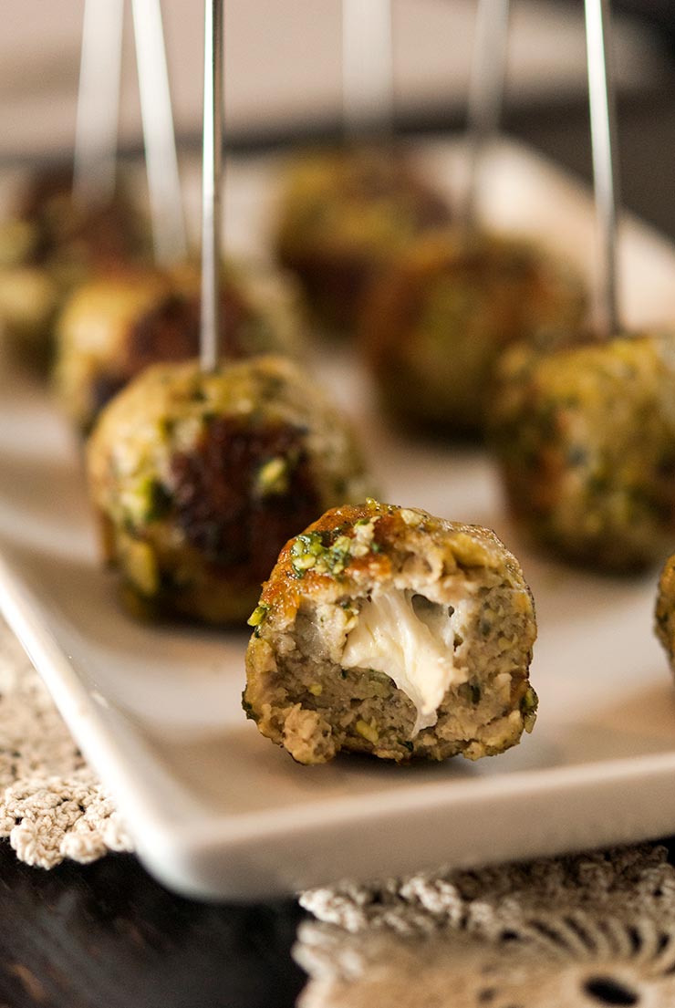 These incredible Italian turkey meatballs with homemade pesto and mozzarella will become everyone’s favorite appetizer at the party! This recipe is so simple and SO delicious! With melted cheese centers and that fresh, Italian pesto flavor, these perfect little appetizers make such an enticing hors d'oeuvre. They’re also gluten-free! #appetizers, #gluten-free, #pesto, #meatballs A sliced meatball on a plate with melted cheese inside, in front of 7 other meatballs.