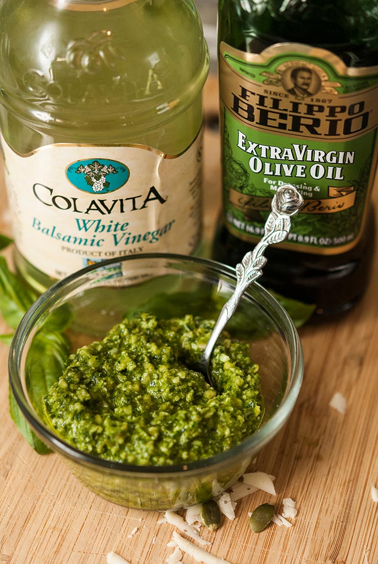 A small bowl of pesto on a wooden board in front of a bottle of balsamic vinegar and olive oil.