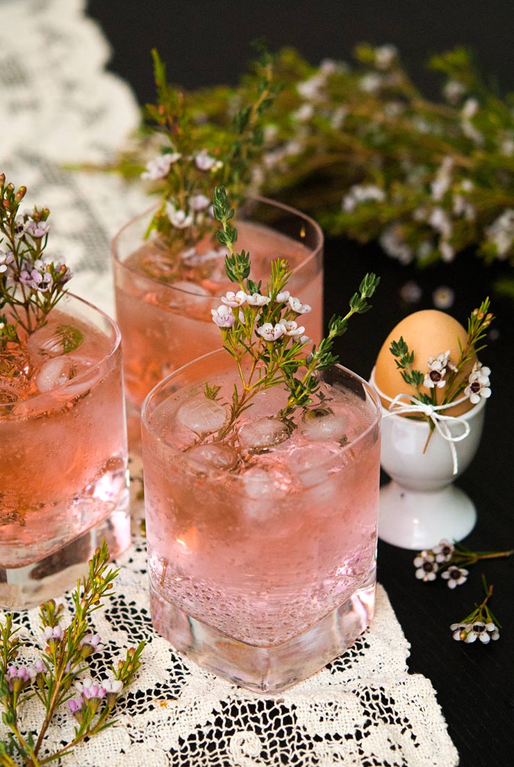 3 pink gin and tonic cocktails on a lace table cloth, garnished with and surrounded by floral greenery and thyme.