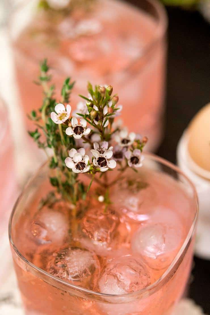 These pink gin and tonics are perfect for a baby or bridal shower, elegant brunch or any time you just want to feel a little lady-like. #cocktails, #babyshower, #bridalshower, #summercocktails, #brunchcocktails, #pink via @shekeepsalovelyhome A closeup of small flowers and thyme in a pink cocktail, in front of 2 de-focused cocktails.