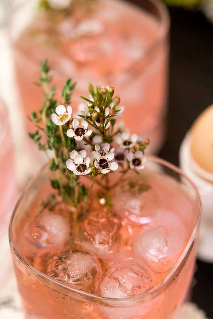 A closeup of small flowers and thyme in a pink cocktail, in front of 2 de-focused cocktails.