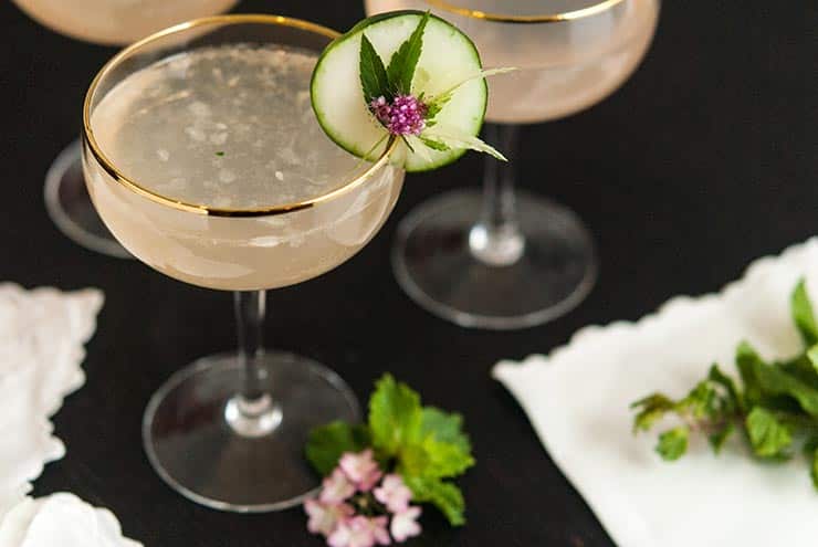 A cocktail with a cucumber and flower garnish on a black table beside 2 other cocktails.