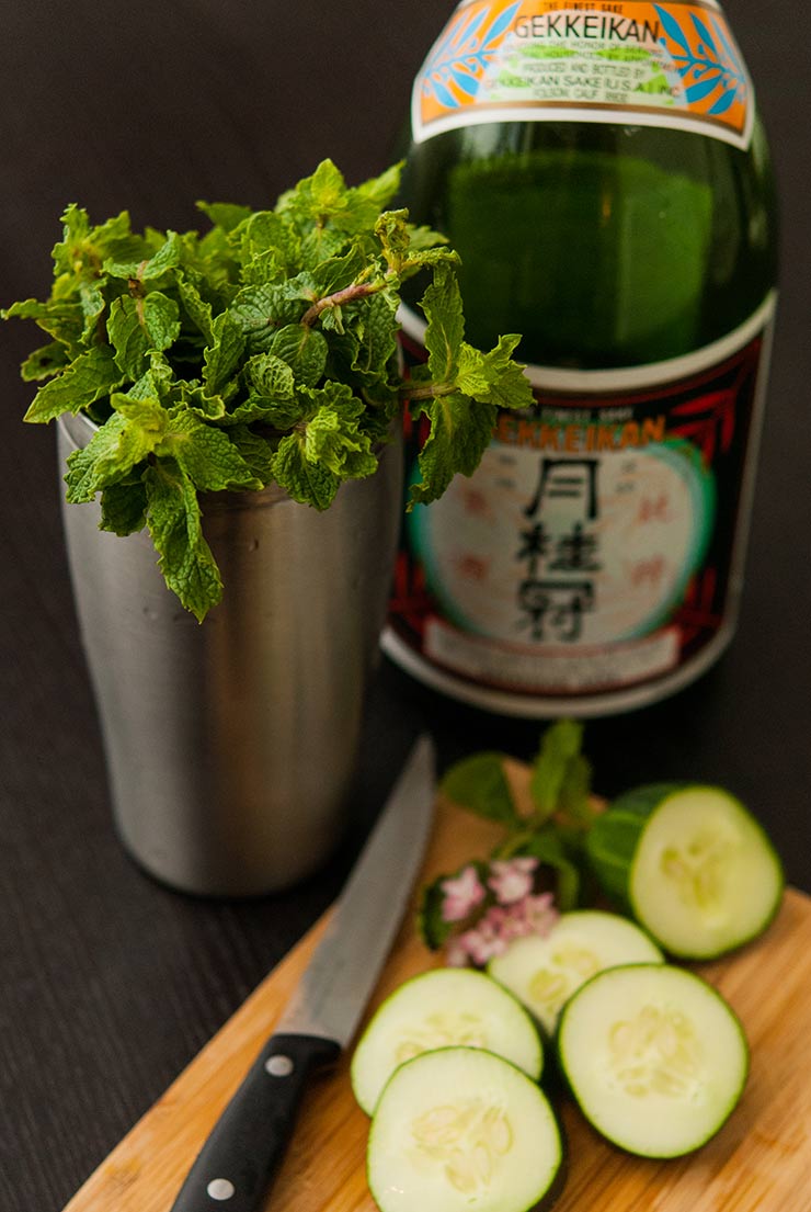 A cocktail shaker full of mint next to a bottle of sake. A cutting board with sliced cucumbers in in front.