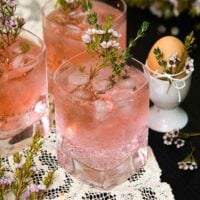 3 pink gin and tonic cocktails on a lace table cloth garnished with and surrounded by floral greenery and thyme, beside an egg in an egg cup.