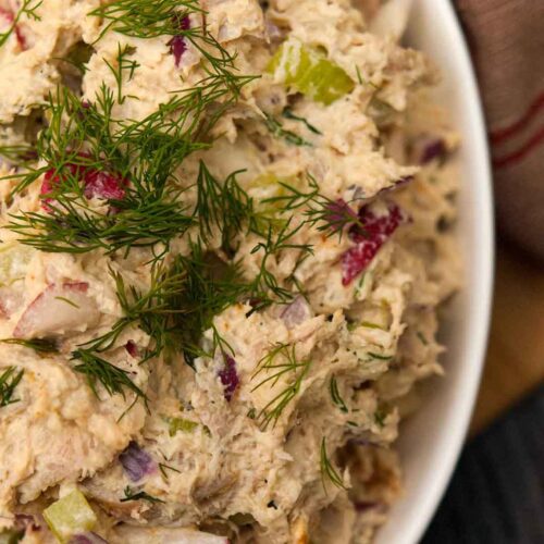 A bowl of chicken salad, topped with dill beside a vintage tea towel.