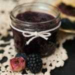 A small jar of jam, tied with a string bow on a lace table cloth, and 2 berries in front of it.