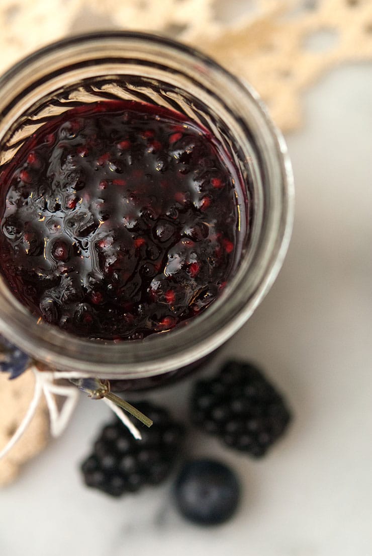 Blueberry jam in a glass jar on top of a marble table with a lace runner. There are 3 berries beside the jar.