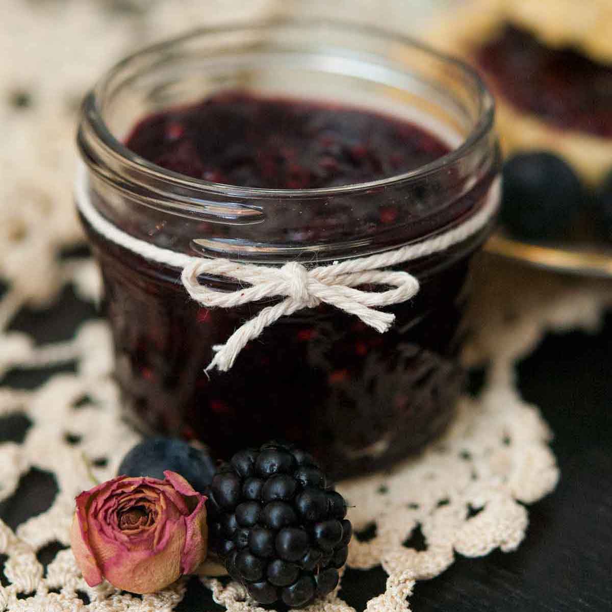 A jar of jam tied with a string with a sprig of lavender tucked into the string with a lace table cloth in the background.