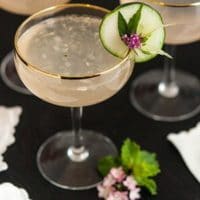 A cocktail garnished with cucumber, a leaf and flower with 2 other glasses in the background and napkins on the table.