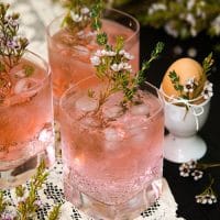 3 pink cocktails, garnished with flowers and rosemary on a lace cloth, beside an egg in an egg cup, tied with flowers.