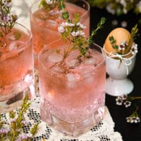 3 pink cocktails, garnished with flowers and rosemary on a lace cloth, beside an egg in an egg cup, tied with flowers.