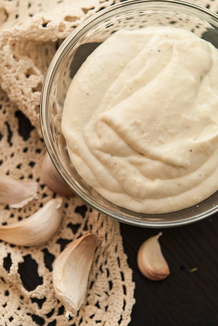 A bowl of roasted garlic sour cream on a table next to a lacy runner and garlic cloves.