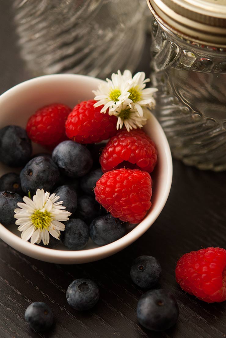 A small white bowl full of raspberries, blueberries and small daisies on a black table.