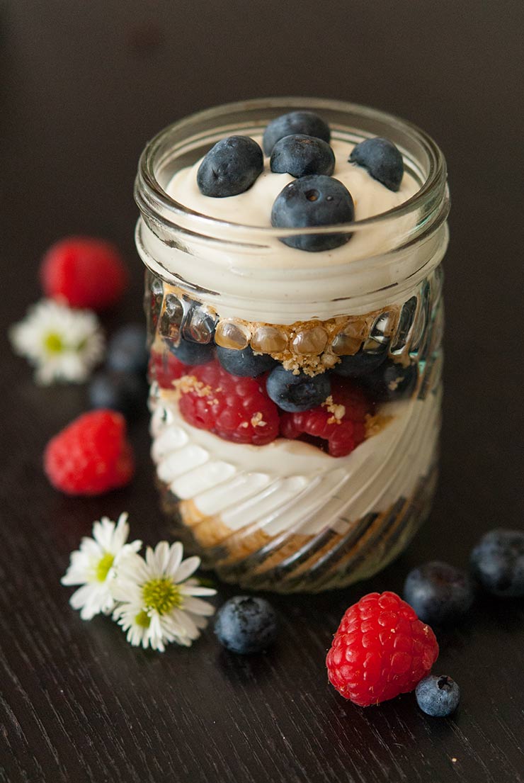 A mason jar with layered cheesecake mouse, cookie crumbles and berries on a black table.