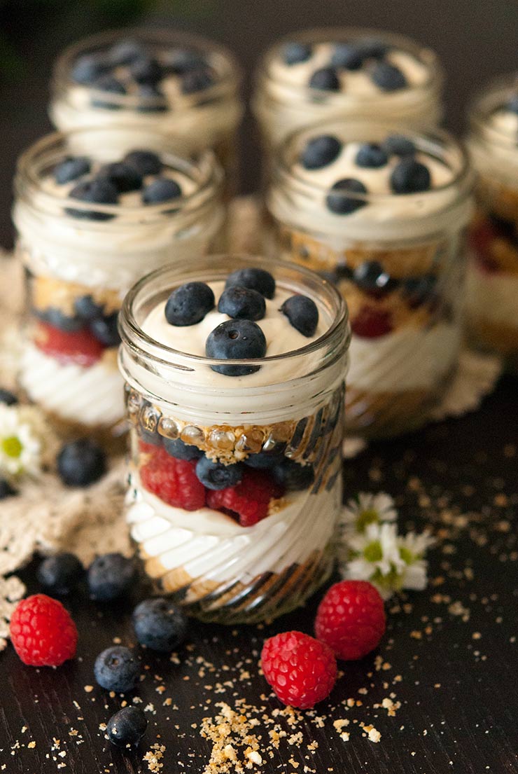 6 mason jars with cheesecake mouse, cookie crumbles and berries on a black table, sprinkled with cookie crumbs and berries.