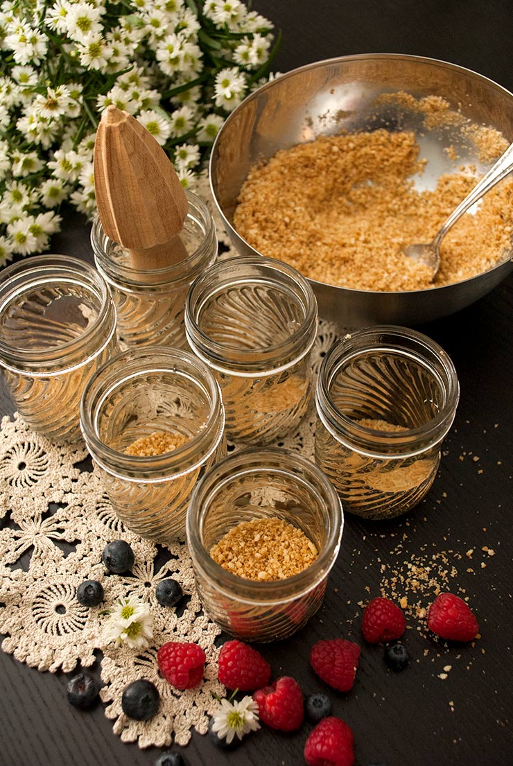 6 mason jars with cookie crumbles on the bottom on top of a lace cloth beside a bowl of cookie crumbles and a bouquet of daisies.