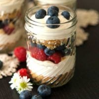 A mason jar with cheesecake mousse, cookie crumbles and berries on a black table, with a few berries and flowers at its base.