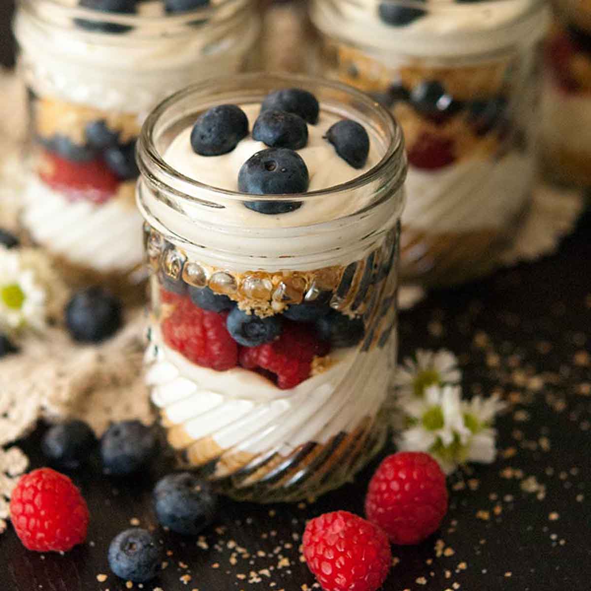 6 mason jars with cheesecake mouse, cookie crumbles and berries on a black table, sprinkled with cookie crumbs and berries.