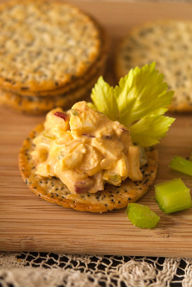 A cracker with sriracha egg salad and a celery leaf in front of small stacks of crackers on a wooden plate.