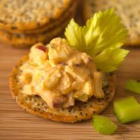 A cracker with sriracha egg salad and a celery leaf in front of small stacks of crackers on a wooden plate.