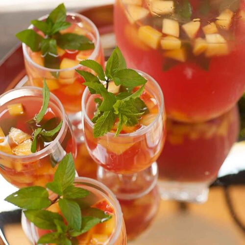 A silver tray with 4 glasses of fruit-filled watermelon sangria, garnished with mint, next to a full pitcher.