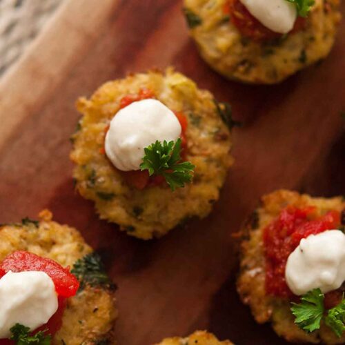 5 cauliflower cakes on a wooden board, topped with marinara, cream, and a little fresh parsley.