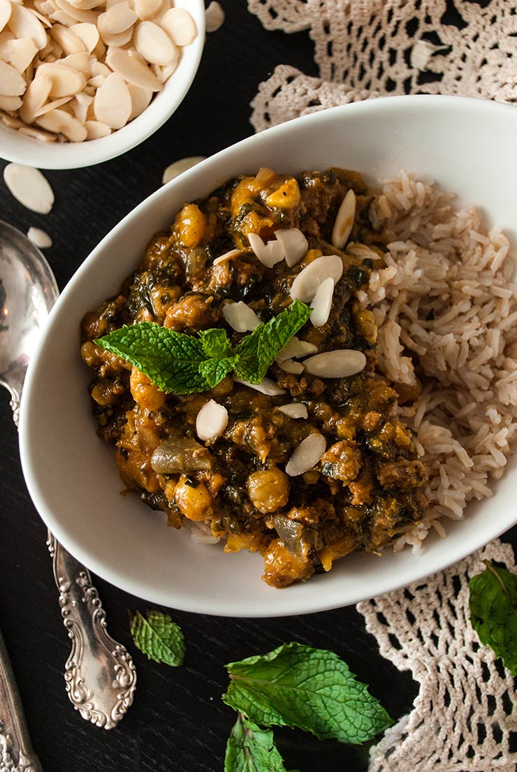 A bowl of curry, turkey butternut squash on a table with a lace table cloth, a bowl of shaved almonds and a few mint leaves.