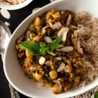 A bowl of curry, turkey butternut squash on a table with a lace table cloth, a bowl of shaved almonds and a few mint leaves.