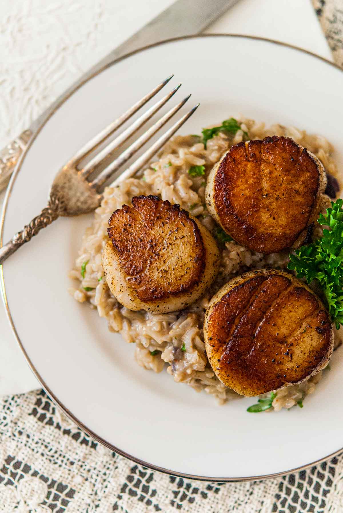 3 seared scallops on top of creamy rice, garnished with a sprig of parsley, on a place beside an antique fork.