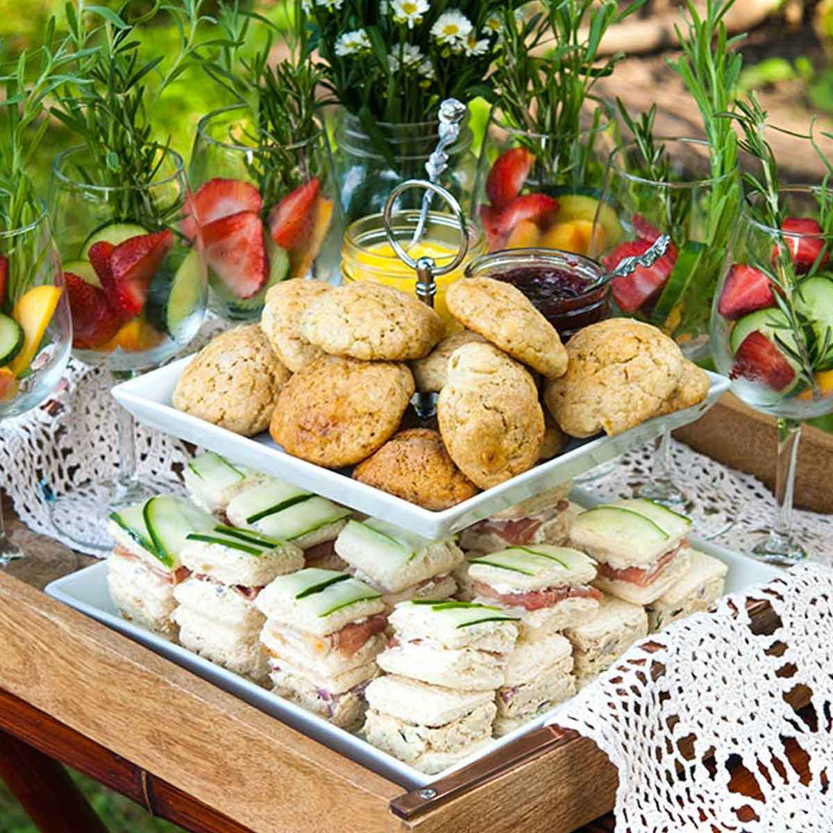 An appetizer tier on a tray with lace, holding scones and cucumber sandwiches, surrounded by cocktail glasses and flowers.