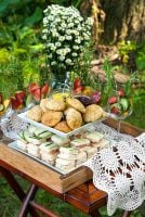 An appetizer tier on a tray with lace, holding scones and cucumber sandwiches, surrounded by cocktail glasses and flowers.