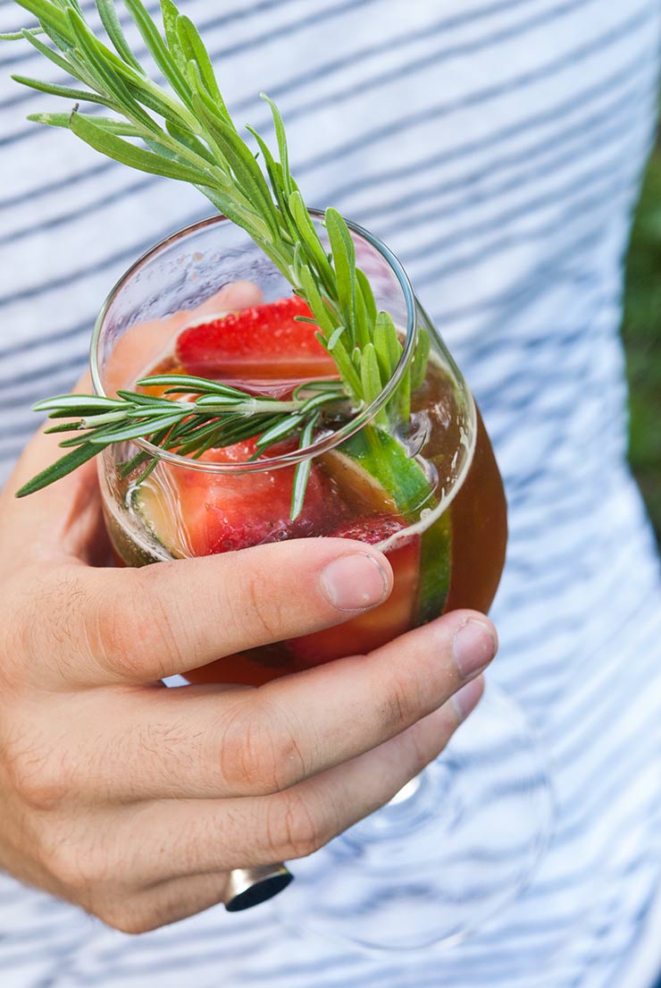 A hand holding a strawberry Pimm’s Cup cocktail with fruit and herb garnishes in front of a striped shirt.