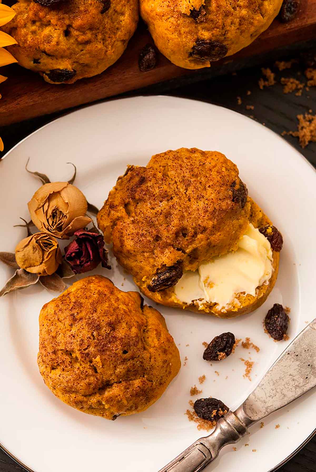 Pumpkin scones on a plate, smeared with butter - the plate is garnished with raisins and a few dry roses.